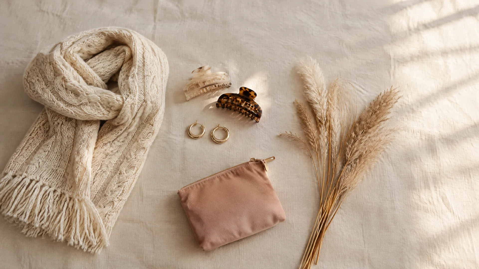 Cream cable scarf, claw clips, gold-tone hoops and a blush pouch beside dried pampas grass