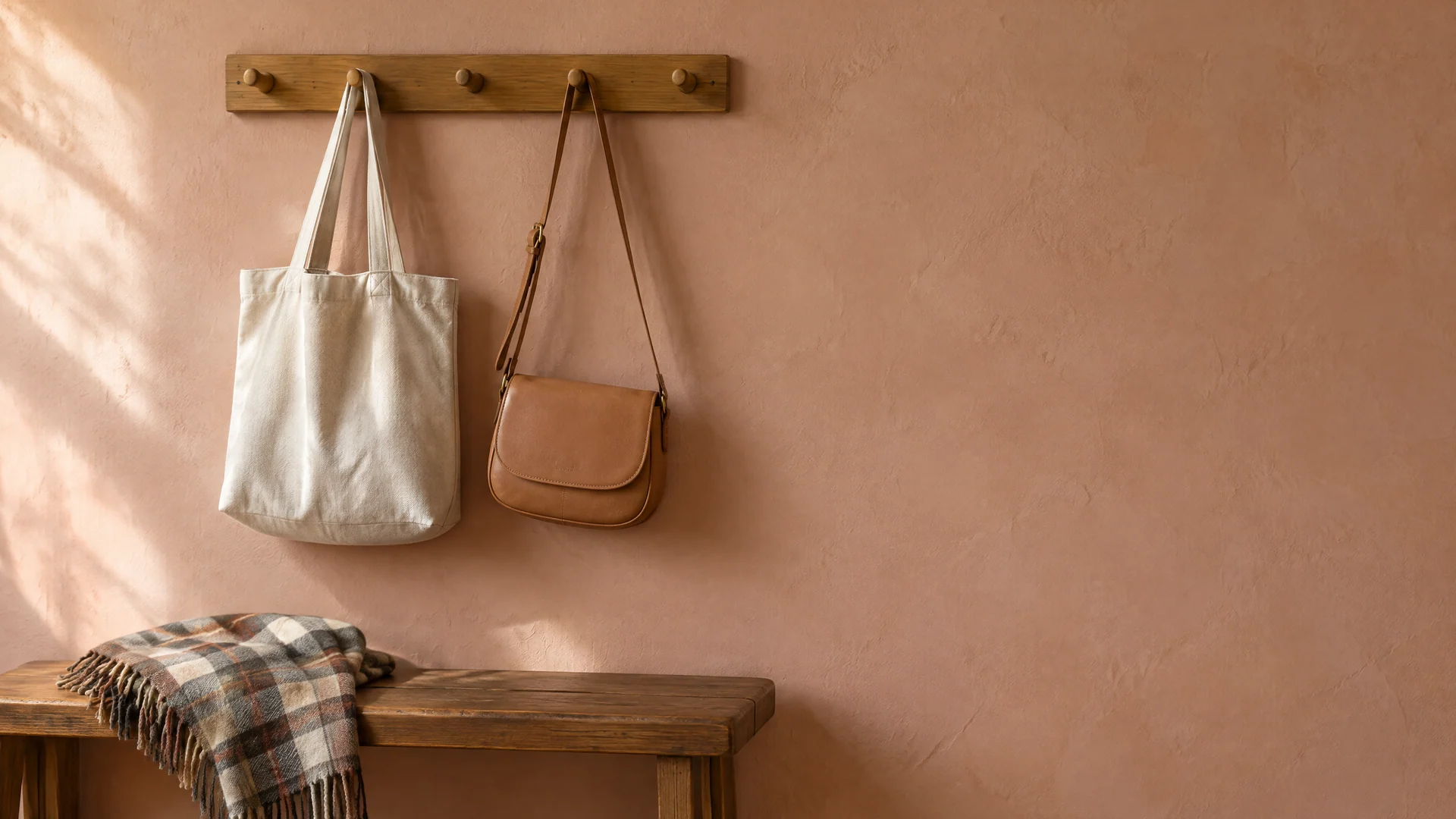 Natural tote and tan crossbody bag hanging from a peg rail above a bench with a plaid scarf